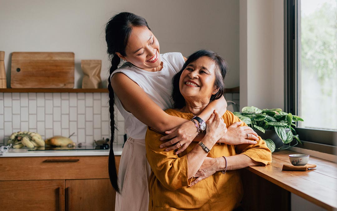 woman hugs her mother while she sits in the kitchen drinking tea