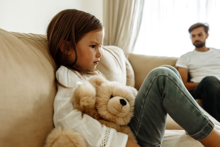 little girl with teddy bear on sofa, man in background